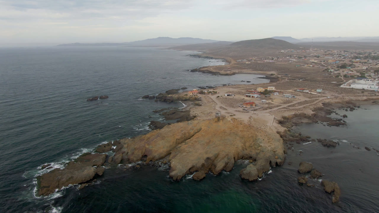 Aerial view over Bahia Ascuncion, Mexico, with dark ocean at left, a tan rocky headland in the foreground, low town buildings inland, distant hills, and no visible people.