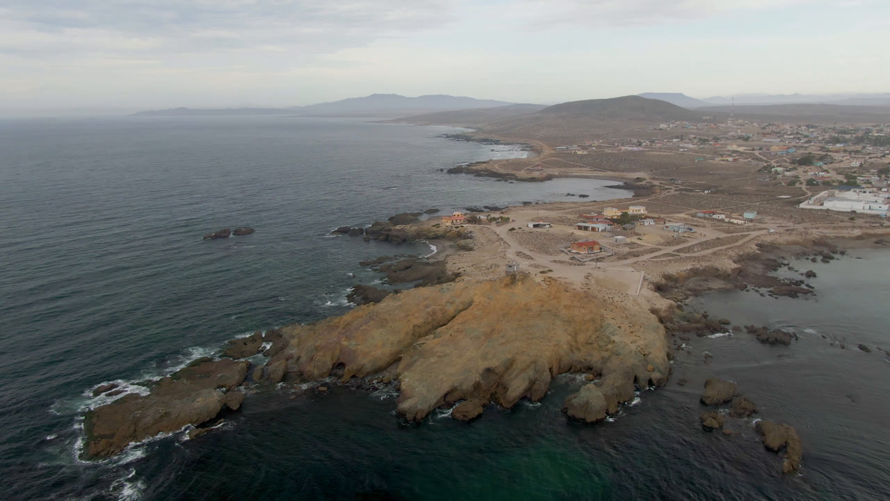 Aerial frame from Bahia Ascuncion showing the ocean filling the left side, breaking surf around dark rocks, a tan point below, inland roads and buildings, and no visible people.
