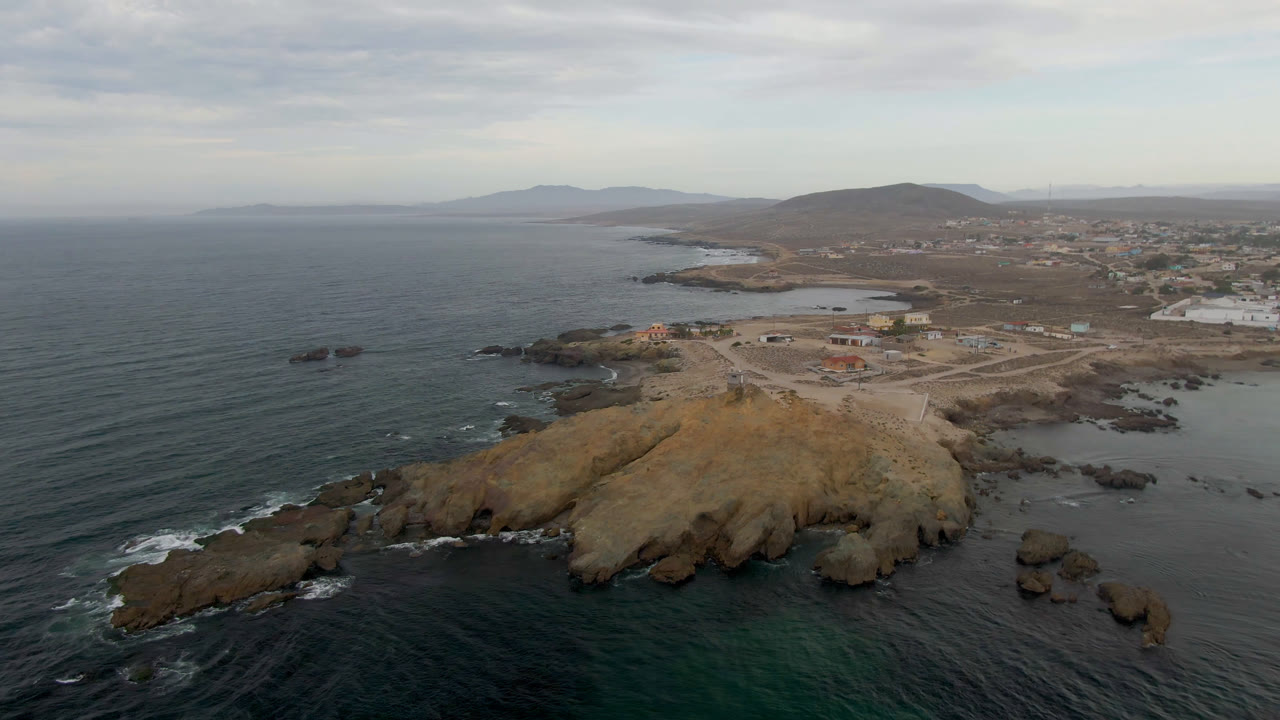 Aerial frame over Bahia Ascuncion, Baja California Sur, showing dark ocean water, a tan rocky point, scattered offshore rocks, distant hills, coastal buildings, and no visible people.