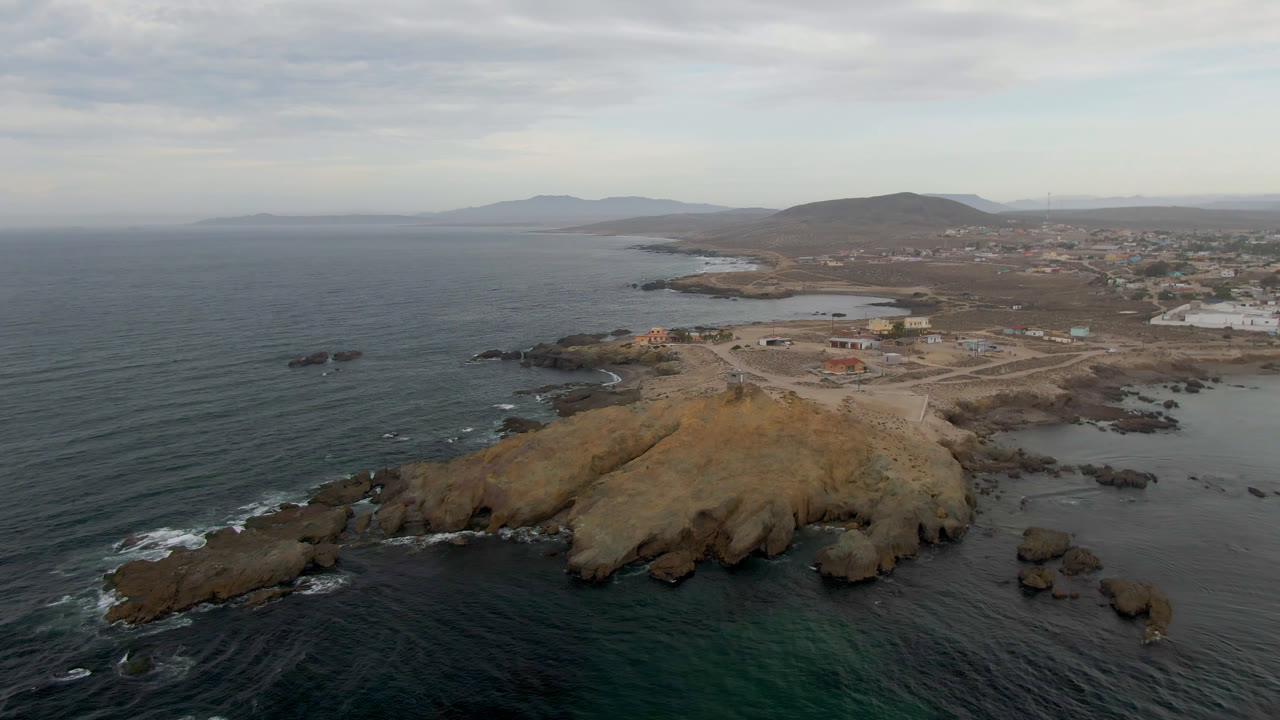 Aerial view in Bahia Ascuncion with the rocky headland centered, dark water and small white wave lines at the rocks, low buildings in the distance, and no visible people.