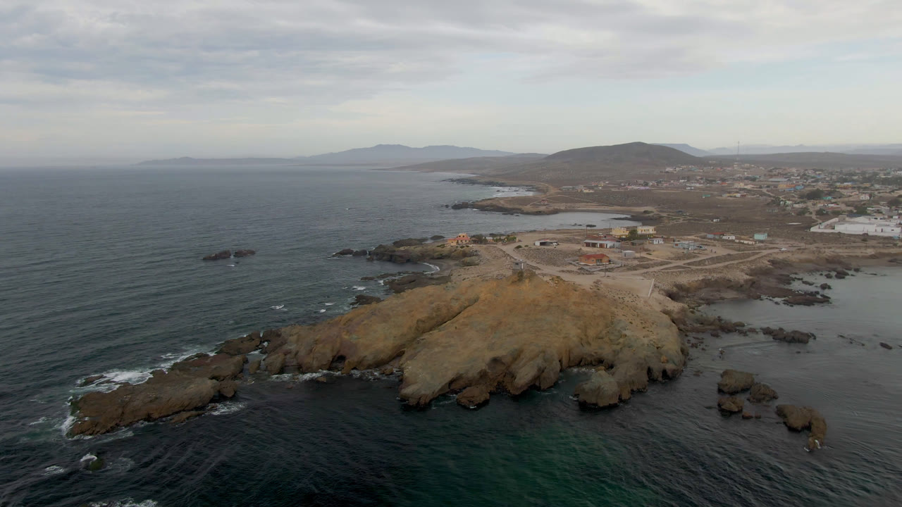 Wide aerial coastline with ocean, rocky outcrop, town edge, and hills.