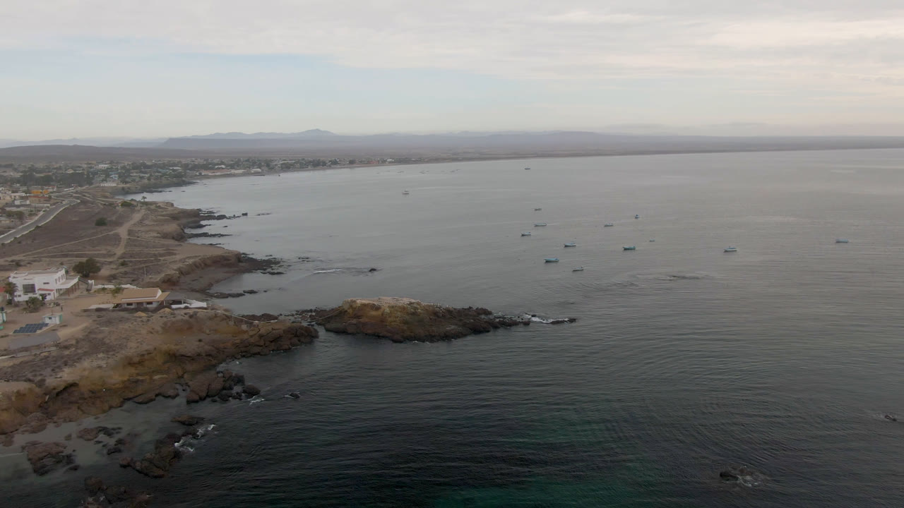 Opening drone frame over Bahia Ascuncion shows small boats on calm water, a rocky point in the foreground, shoreline buildings to the left, distant landforms, and no visible people.