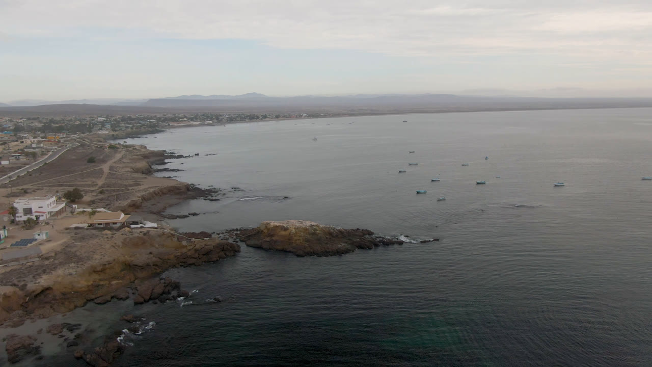 At 0:01, the Bahia Ascuncion aerial keeps the rocky foreground and low shoreline buildings visible while more anchored boats sit across the gray-blue bay, with no visible people.