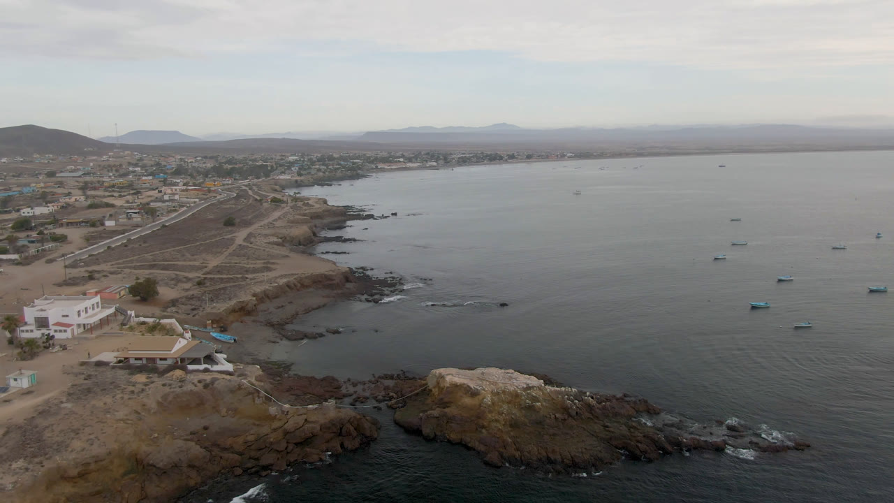 Aerial view over Bahia Ascuncion, Baja California Sur, with anchored boats on gray-blue water, rocky shoreline, low buildings, dirt tracks, distant hills, and no visible people.