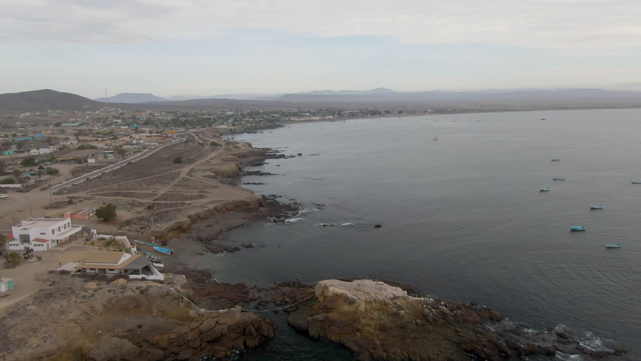 Wide aerial frame in Bahia Ascuncion showing small boats spread across calm bay water, a rocky point in the foreground, low shore buildings, dirt paths, and no visible people.