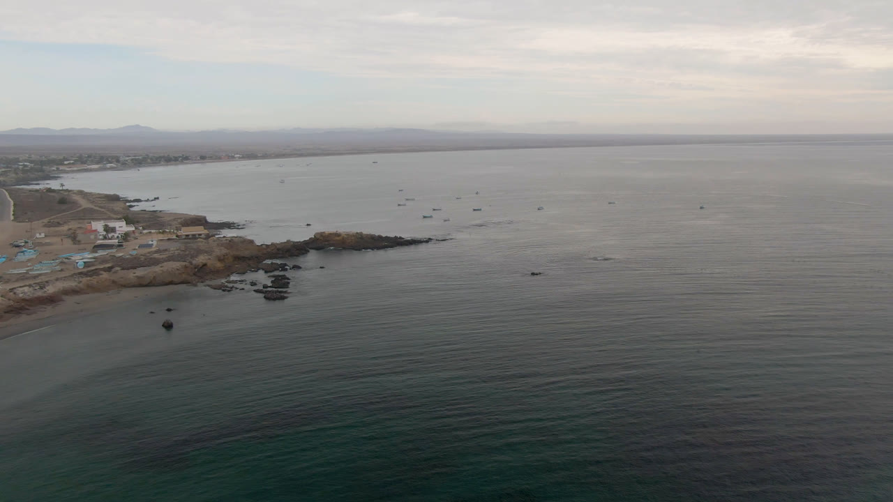 Aerial view over Bahia Ascuncion water with small boats offshore, a rocky point and light buildings at left, distant hills on the horizon, and no visible people.