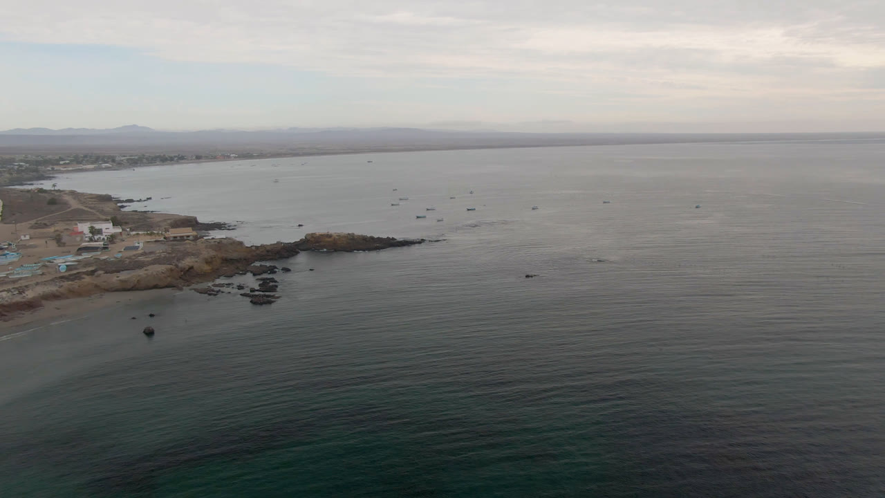 Drone view in Bahia Ascuncion looking across dark green water toward scattered boats, a low rocky shoreline at left, pale buildings near shore, and no visible people.