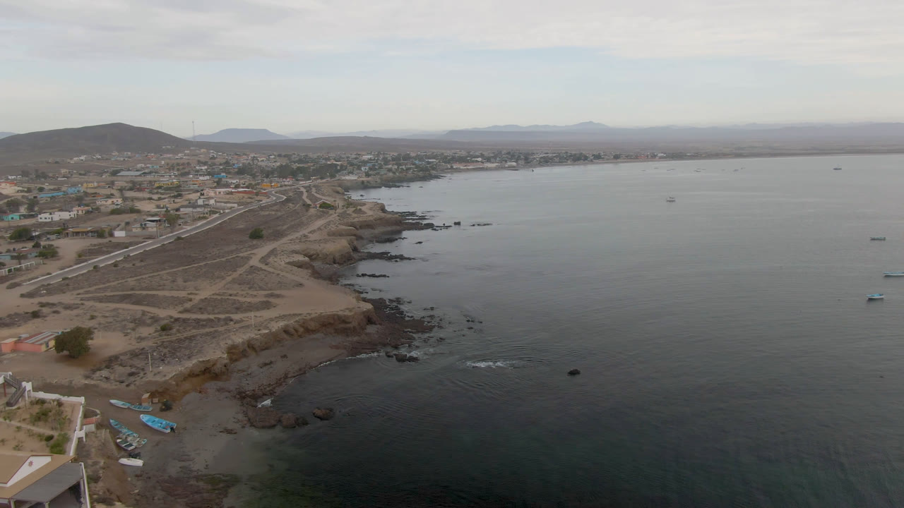 Aerial frame over Bahia Ascuncion showing dark bay water beside a rocky shoreline, dirt tracks, small boats near the beach, low town buildings, distant hills, and no visible people.