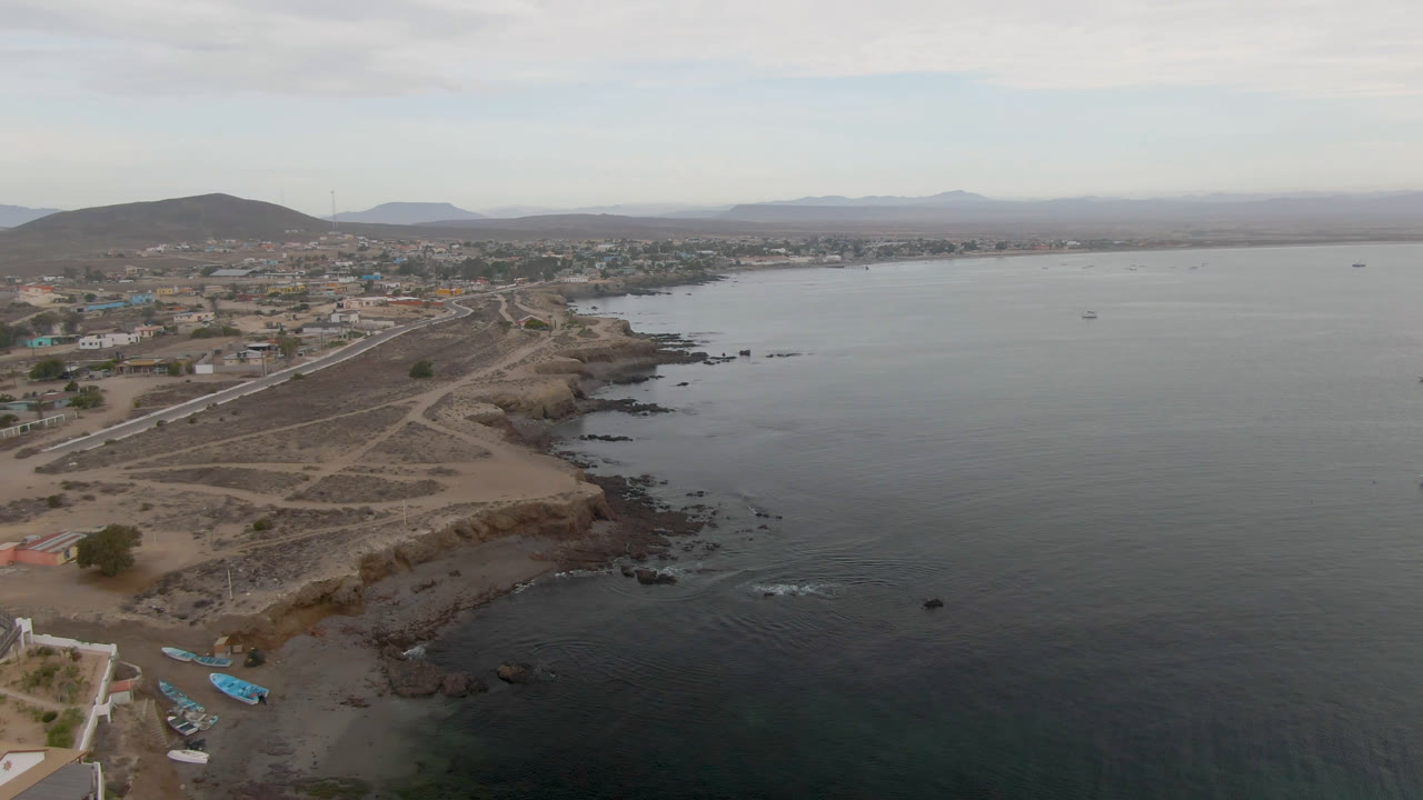Drone view over Bahia Ascuncion with a coastal road and dirt paths leading along the cliff edge, town blocks inland, a broad bay to the right, hazy hills on the horizon, and no visible people.