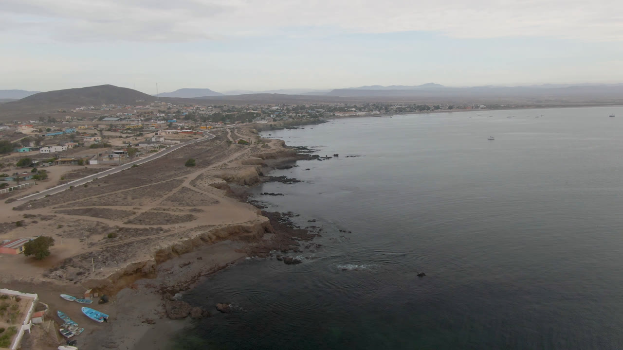 Wide aerial of bay water, rugged shoreline, small town, and low hills under clouds