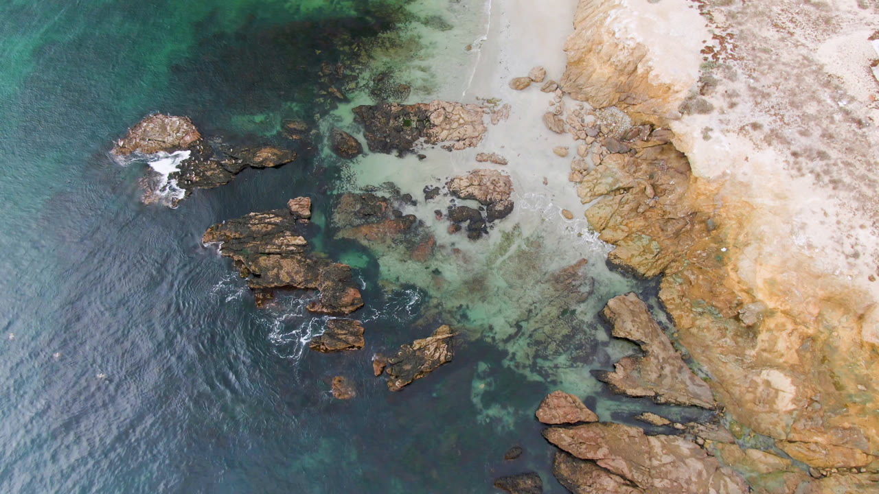 Overhead view of Bahia Ascuncion rocky shoreline with teal water on the left, pale sand and tan rock ledges on the right, and no visible people.