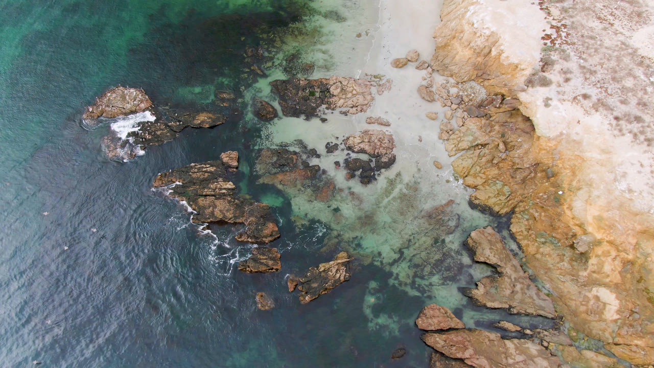 Aerial frame over Bahia Ascuncion showing offshore brown rocks, small white wave breaks, pale sand, and no visible people.