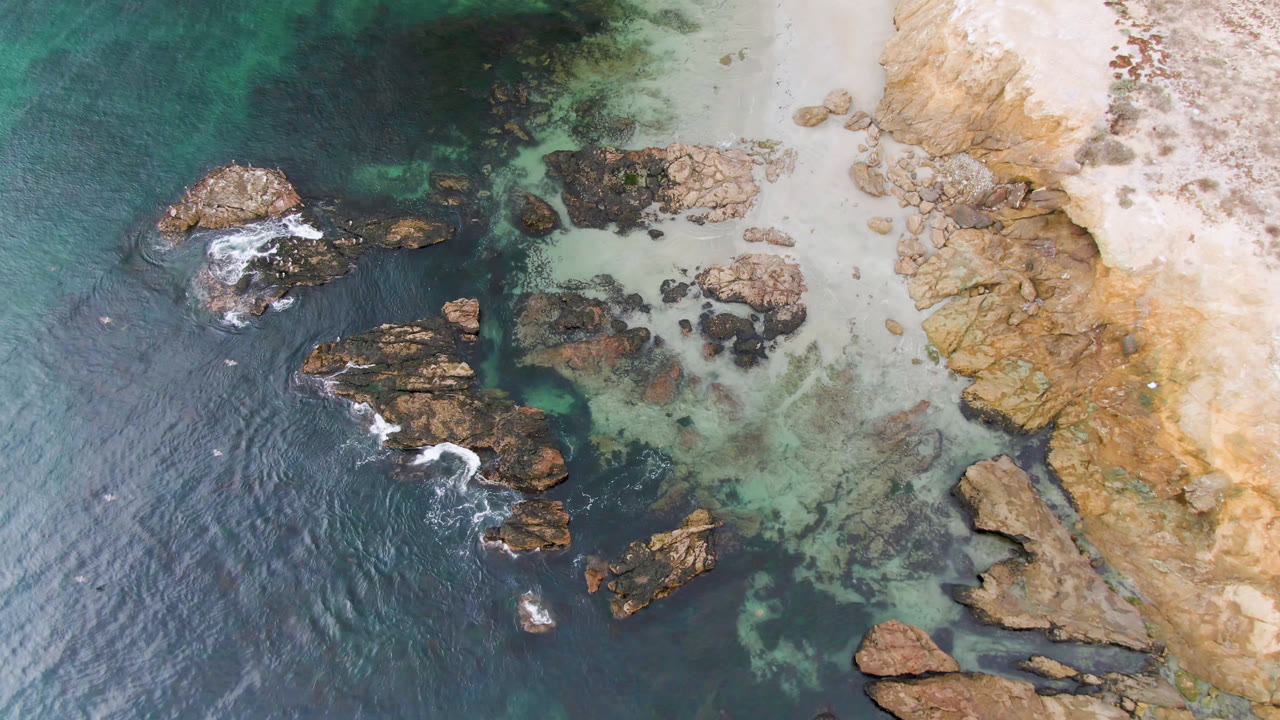 Top-down coast with scattered rocks, shallow green water, and dry bluff edge.