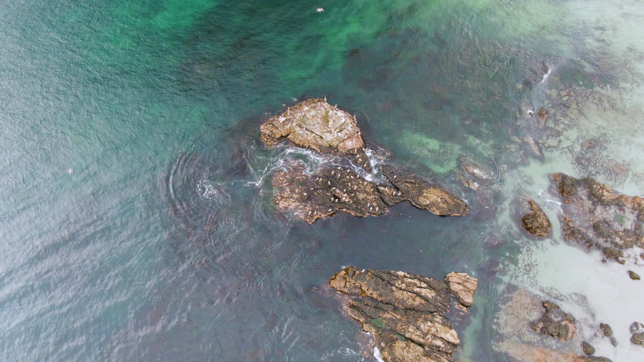 At 0:00, an overhead view in Bahia Ascuncion shows tan rocks surrounded by blue-green shallows and pale sand at the right edge, with no visible people.