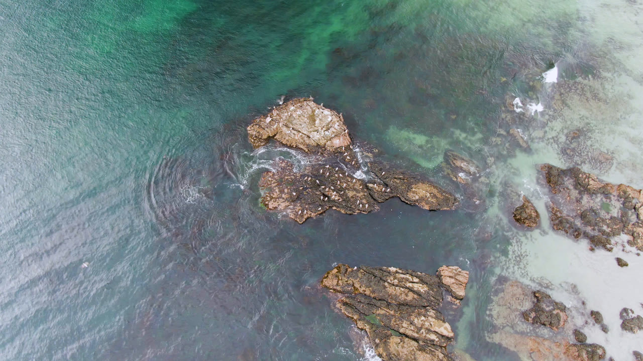 At 0:01, the Bahia Ascuncion frame keeps the rock cluster centered while a small patch of white water appears near the upper right shallows, with no visible people.
