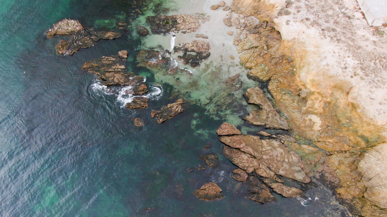 Overhead frame of Bahia Ascuncion coastline with dark teal water on the left, jagged rocks near shore, pale beach and tan cliff on the right, and no visible people.