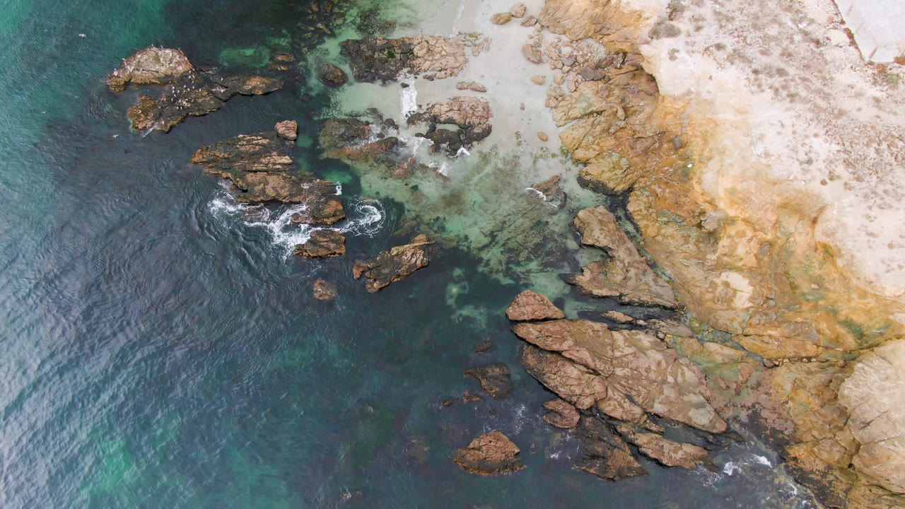 Aerial view over Bahia Ascuncion with offshore rocks surrounded by dark water, clear shallows near the beach, tan cliff rock to the right, and no visible people.