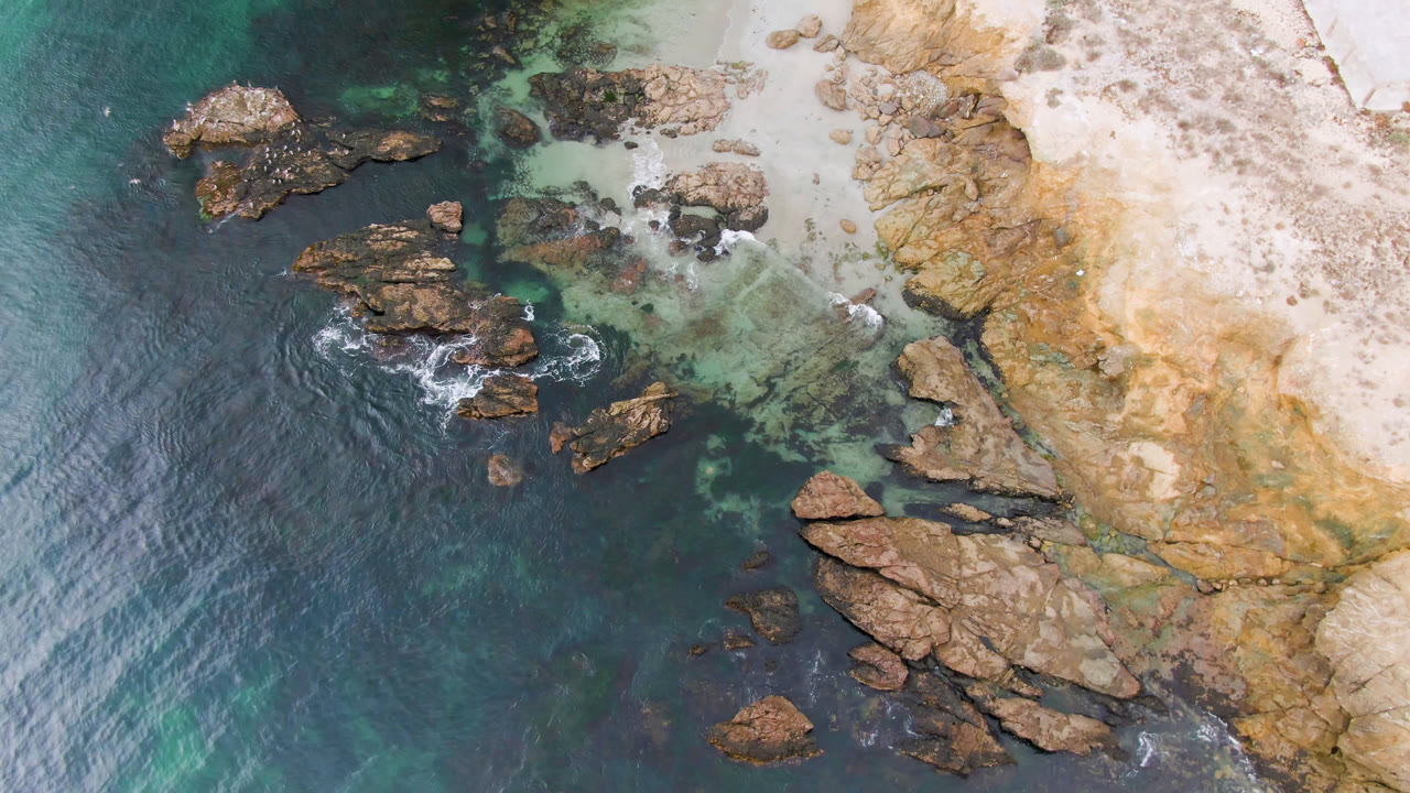 Top-down coastal frame with rocks, turquoise shallows, surf, beach, and cliff.