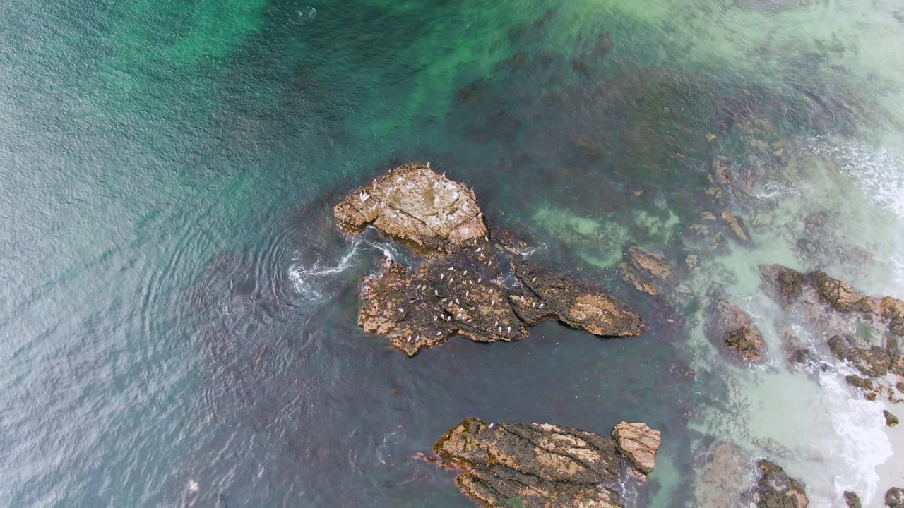 Aerial view over Bahia Ascuncion showing tan rocks in teal and dark blue shallow water, with pale surf along the right edge and no visible people.