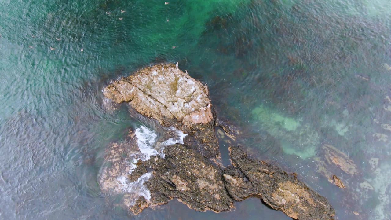 Top-down aerial view in Bahia Ascuncion, Baja California Sur, showing turquoise seawater, dark submerged patches, tan rocks, and no visible people at the start of the clip.