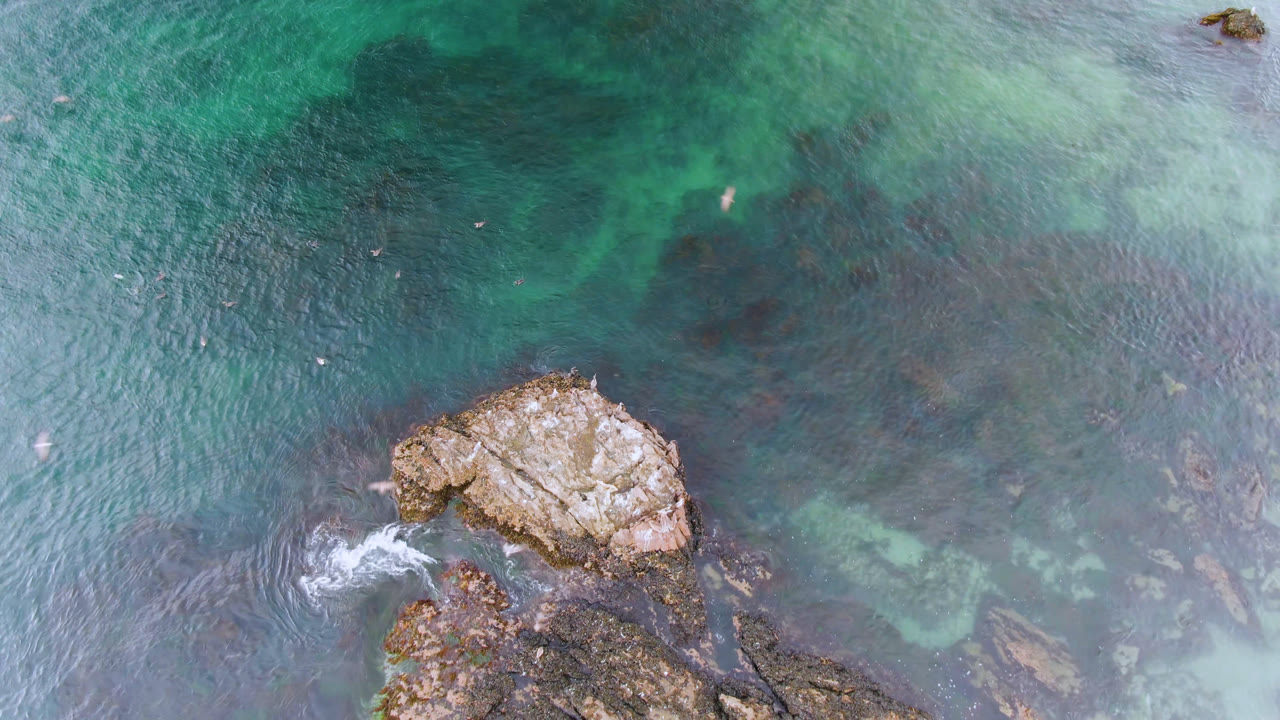Overhead view in Bahia Ascuncion, Baja California Sur, showing turquoise water, exposed rocks, and tiny people near a surfboard at the start of the clip.