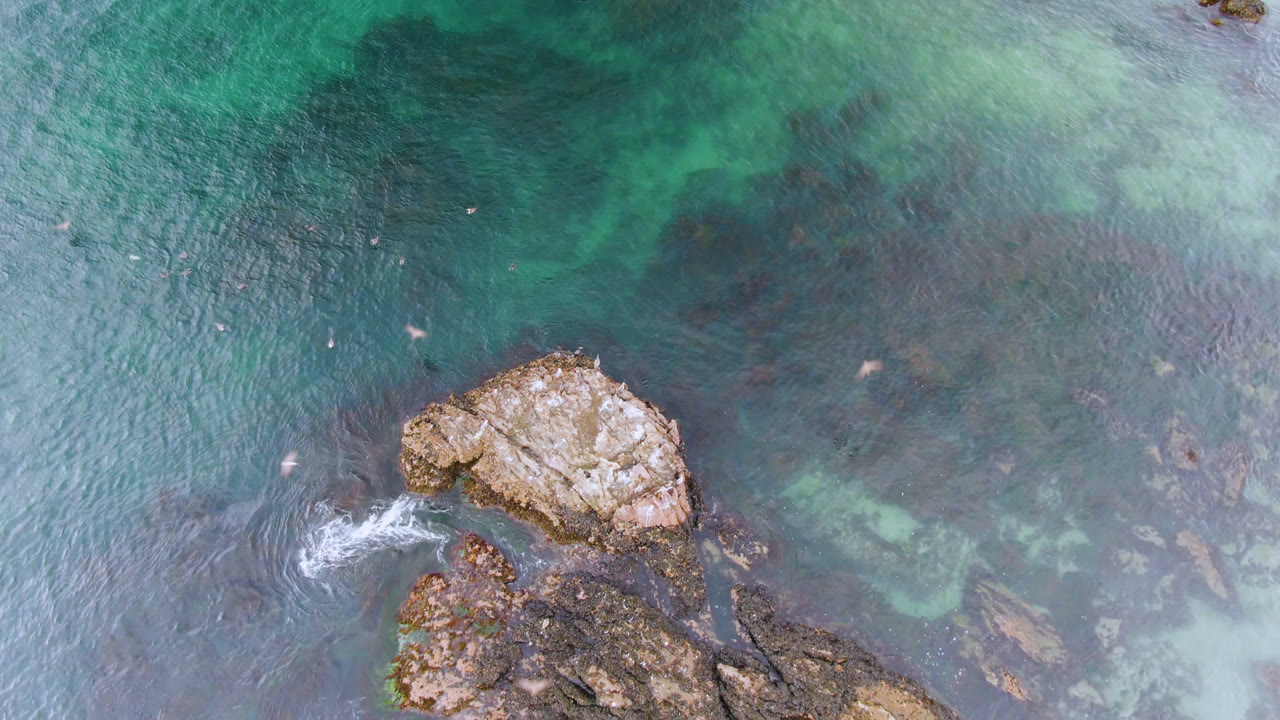 Aerial frame over Bahia Ascuncion water with the main rock cluster lower in frame, pale surf foam at left, and tiny surfboard figures crossing open water.