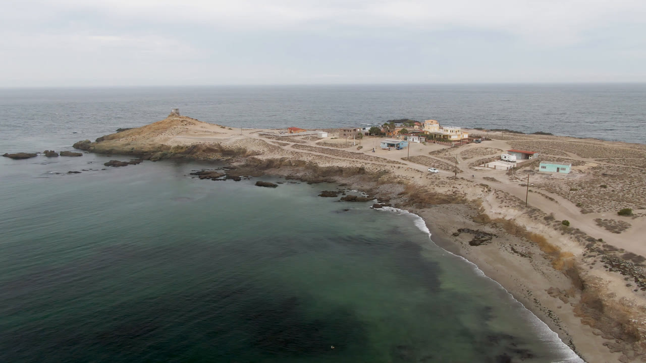 Opening aerial frame over Bahia Ascuncion, Baja California Sur, Mexico, showing green-blue water, a sandy cove, rocky island terrain, small buildings, a lighthouse on the low hill, and no visible people.