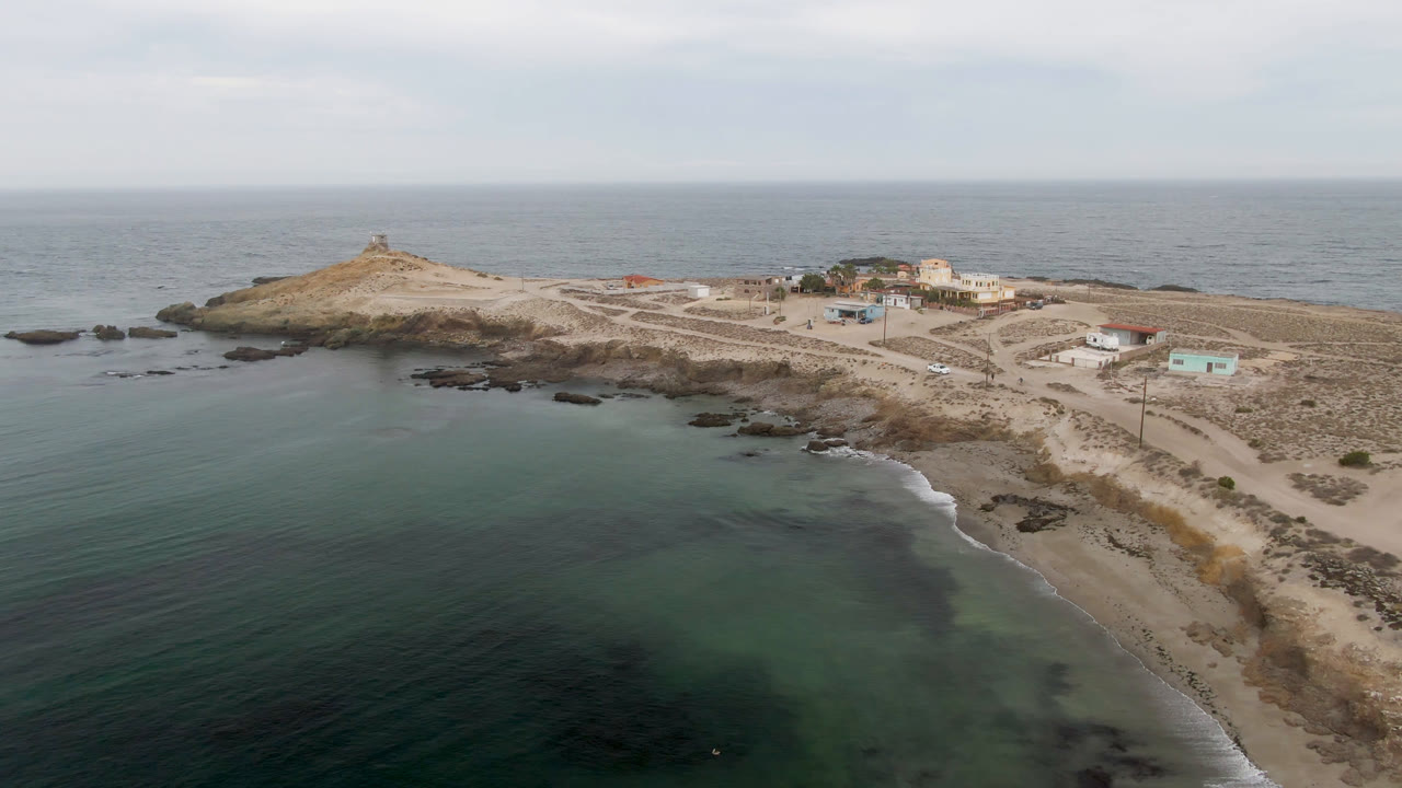 One second into the clip, the Bahia Ascuncion aerial holds on calm nearshore water, a curved sandy beach, rocky edges, pale roads, small structures, the lighthouse hill, and no visible people.