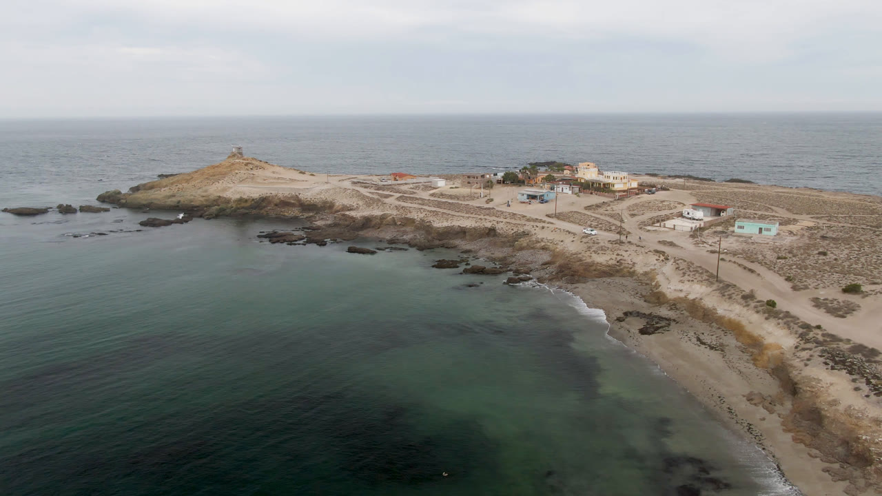 Drone frame of island shoreline with dark rocks, pale roads, buildings, and ocean horizon