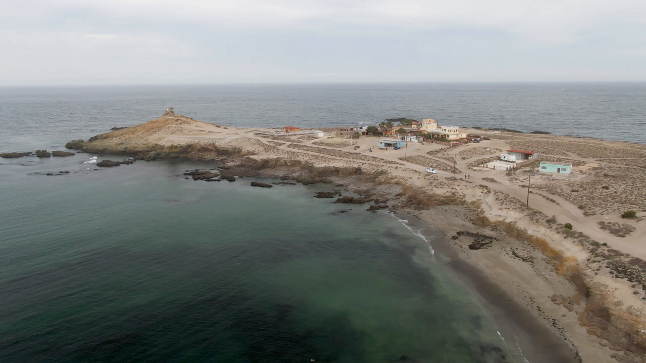 Aerial view of green-blue water, sandy beach, rocky island, buildings, and a small lighthouse rise.