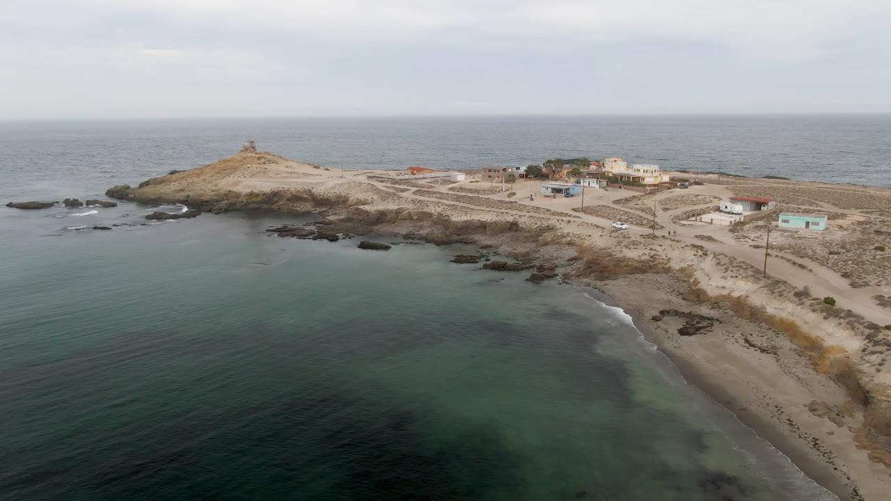 Aerial frame over Bahia Ascuncion showing shallow green-blue water, a narrow sandy beach, dark shoreline rocks, low coastal buildings, and no visible people.