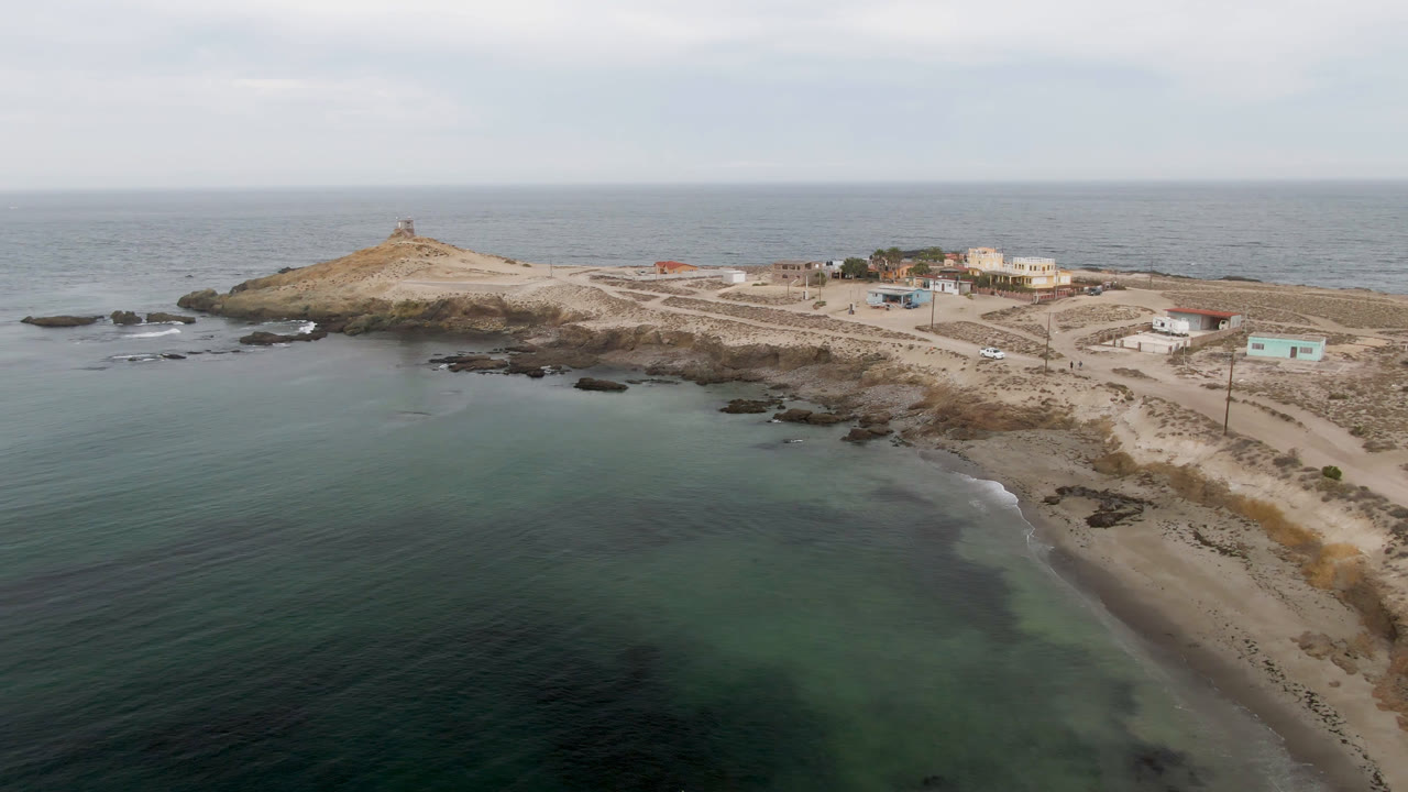 Drone-height view of Bahia Ascuncion with shallow water filling the foreground, the beach running along the right edge, rocky outcrops near the point, and no visible people.
