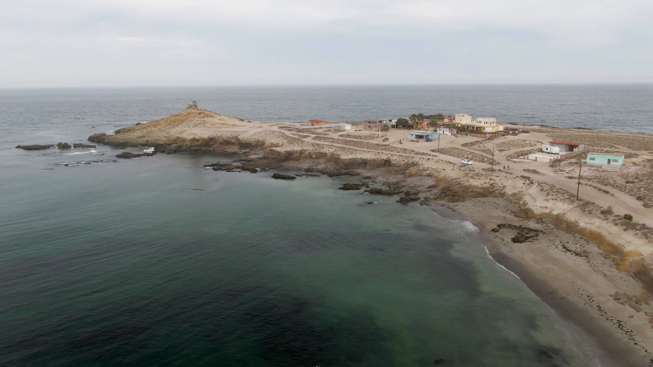 Wide aerial coastline with ocean foreground, sandy shore, rocky point, buildings, and a small structure on the hill.