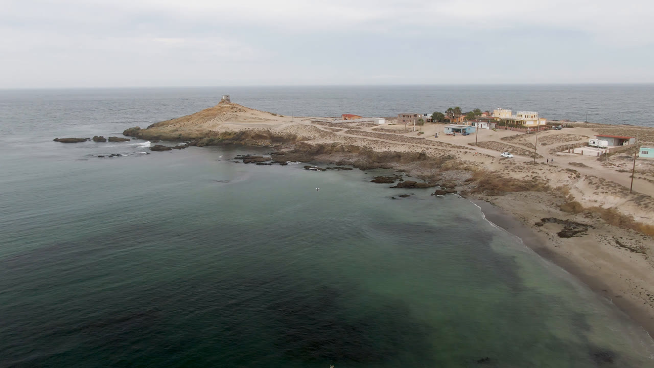 Aerial frame over Bahia Ascuncion shows green-blue water, a rocky sandy island with a small lighthouse on top, coastal buildings to the right, and no visible people.
