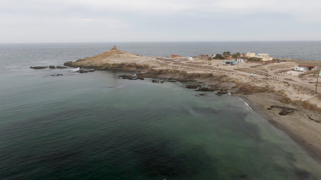The second frame keeps the Bahia Ascuncion lighthouse island near the upper left, with dark rocks along the shore, pale sand, coastal structures, and no visible people.