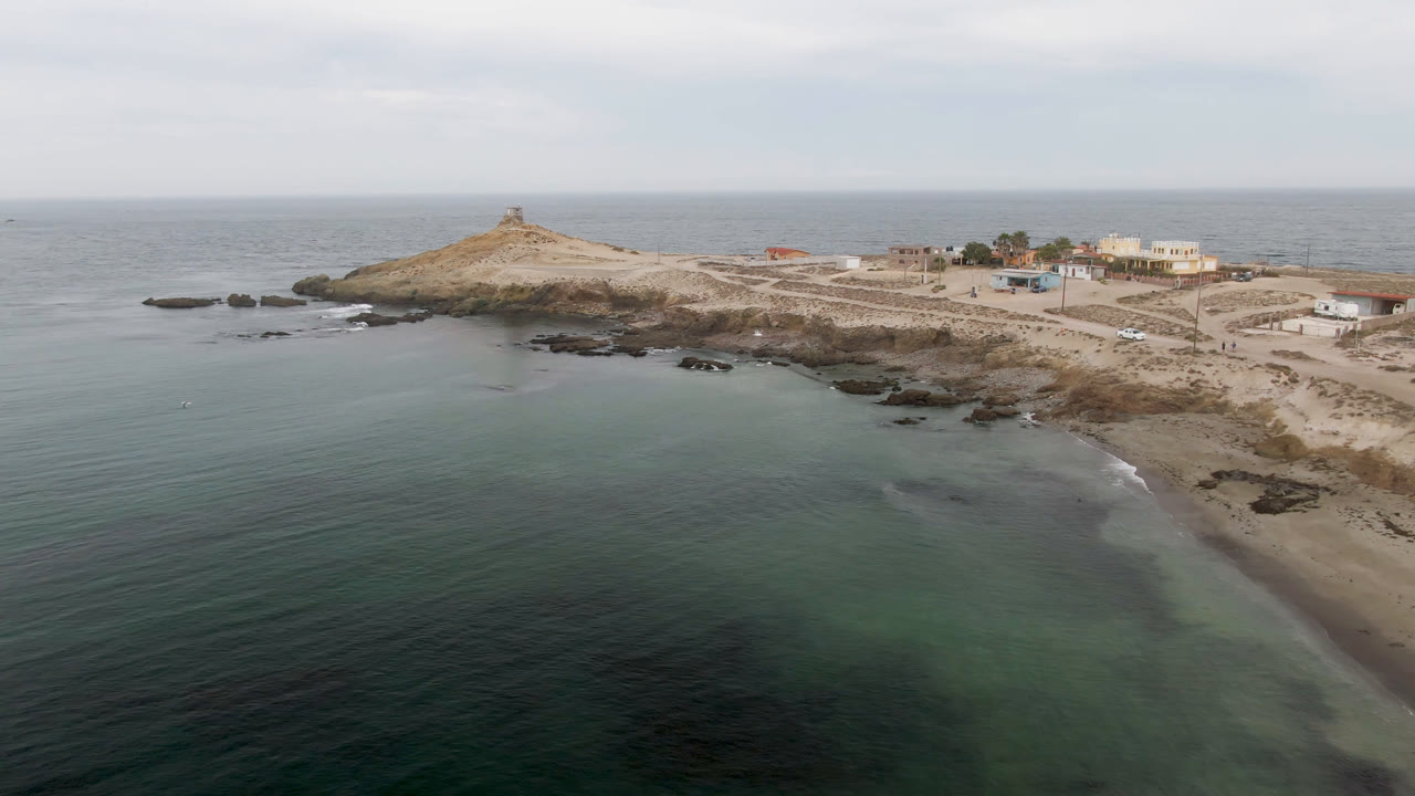 Wide aerial of turquoise water curving around a rocky beach and lighthouse island.