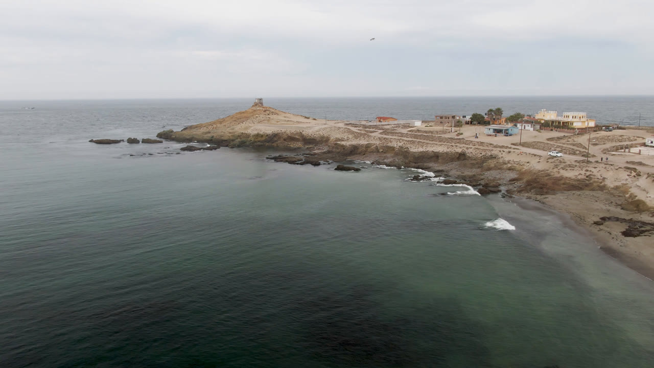 Aerial view over green-blue water toward Bahia Ascuncion shoreline, with a low headland, small lighthouse-like tower, rocks near the surf, coastal buildings to the right, and no visible people.