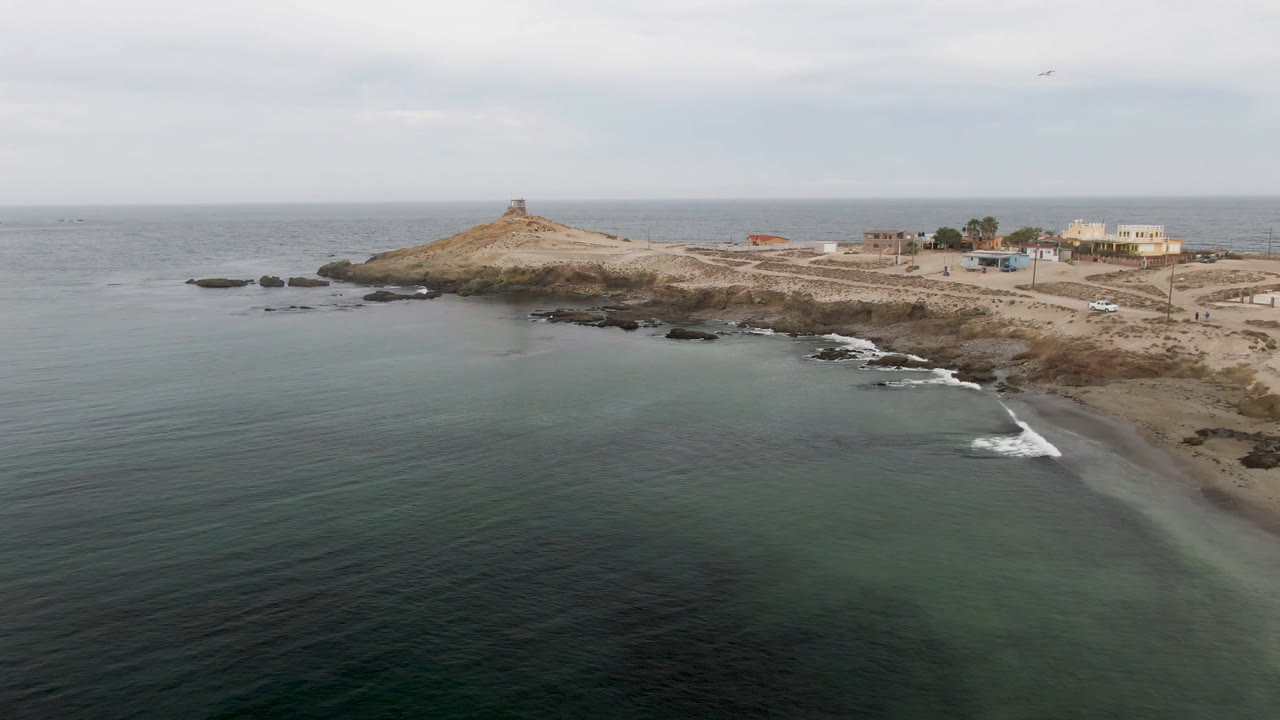 Aerial frame from Bahia Ascuncion showing calm ocean in the foreground, pale surf sliding onto the beach, dark rocks offshore, the lighthouse-like tower on the headland, and no visible people.