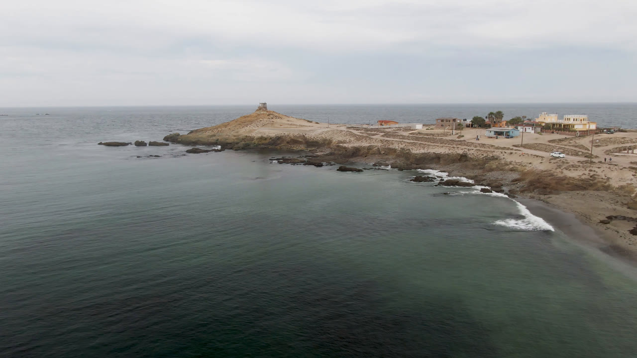 Drone view of open water and rocky beach with a lighthouse point and buildings in the distance.
