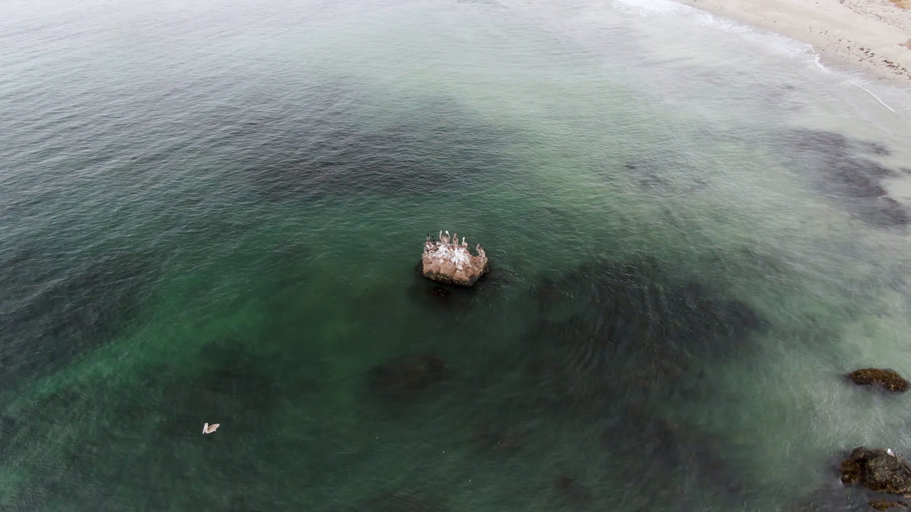 Aerial frame from Bahia Ascuncion showing green coastal water, a rocky outcrop near center-left, a small boat near the lower left, pale surf at the top, and no visible people.