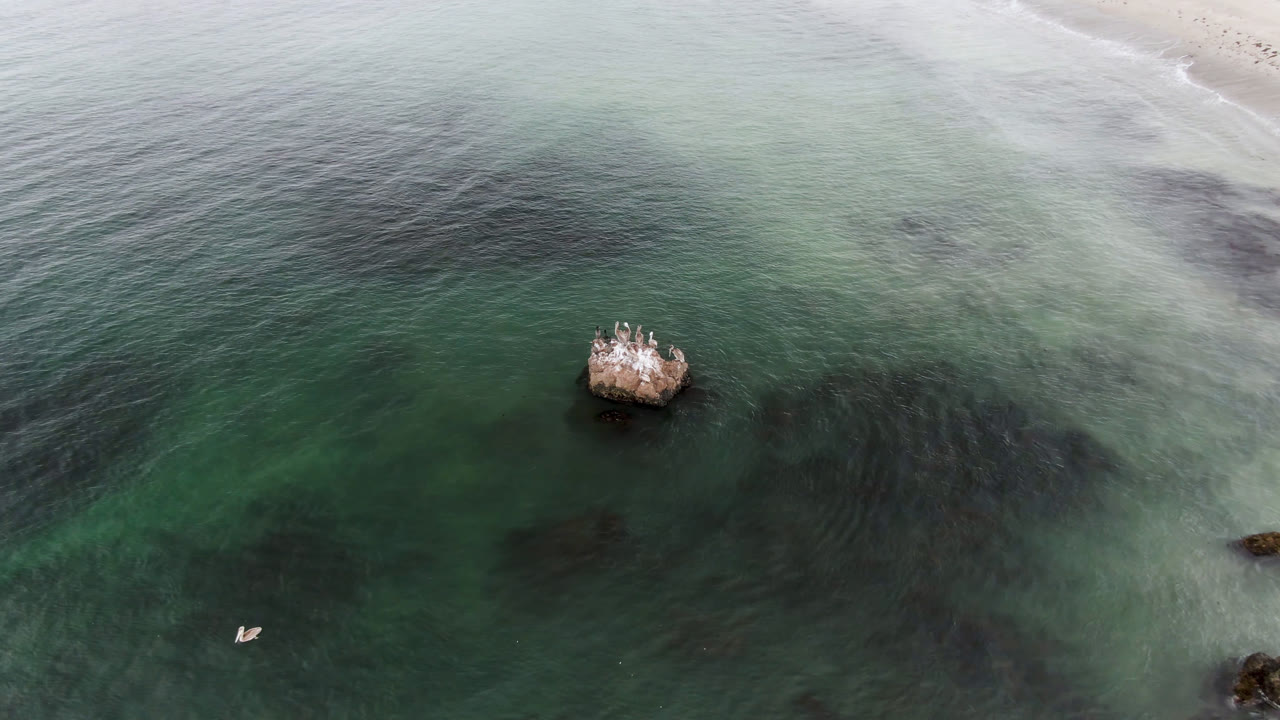 Drone frame from Bahia Ascuncion with the small boat lower in the image, the rocky outcrop left of center, darker underwater shapes, pale beach surf along the top edge, and no visible people.
