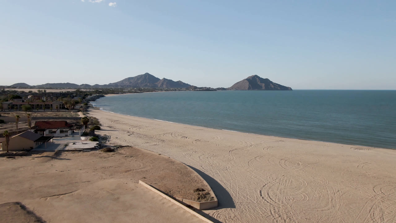 Opening aerial frame over San Felipe BC, Mexico with pale sand below, ocean at right, coastal homes at left, distant mountains on the horizon, and no visible people.