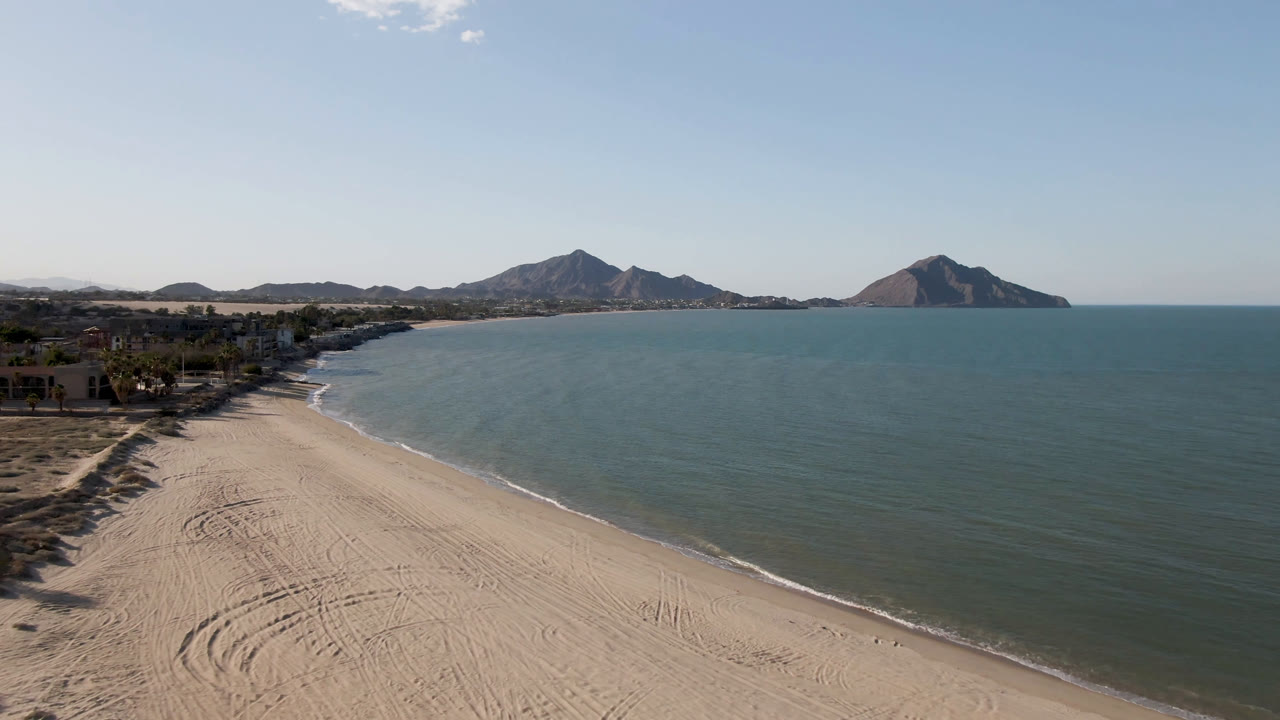 Aerial view over San Felipe, Baja California, Mexico, with a sandy beach in the foreground, blue-green water to the right, distant mountains on the horizon, and no visible people.