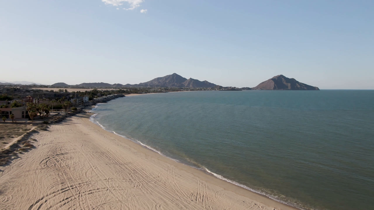 Wide aerial frame from San Felipe, Baja California, Mexico, showing the shoreline curving into the distance, water filling the right side, mountains beyond the bay, and no visible people.