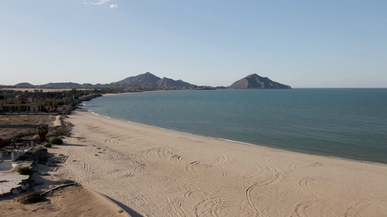 Wide aerial frame of San Felipe, Mexico showing the beach curving along the water, a strip of low coastal buildings at left, distant mountains, and no visible people.