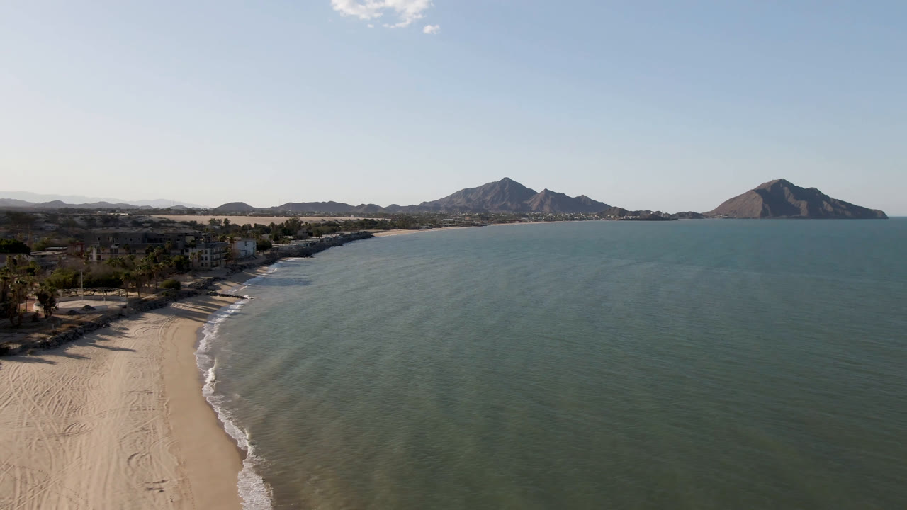 Aerial San Felipe view with the curving sand line closer to frame left, shallow surf tracing the shore, broad ocean water, distant mountains, and no visible people.