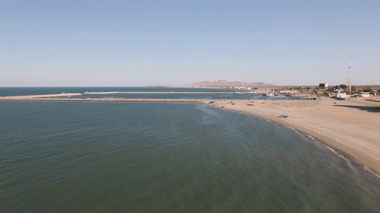 Aerial frame over San Felipe, Baja California, Mexico showing green-blue water in front, a sandy beach and long pier beyond, distant mountains, and no visible people.