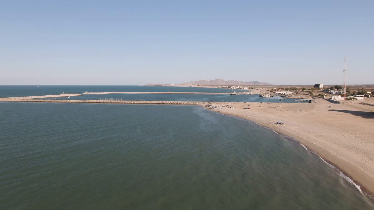 Aerial coastline with calm water, beach, pier, buildings, and a tall mast.