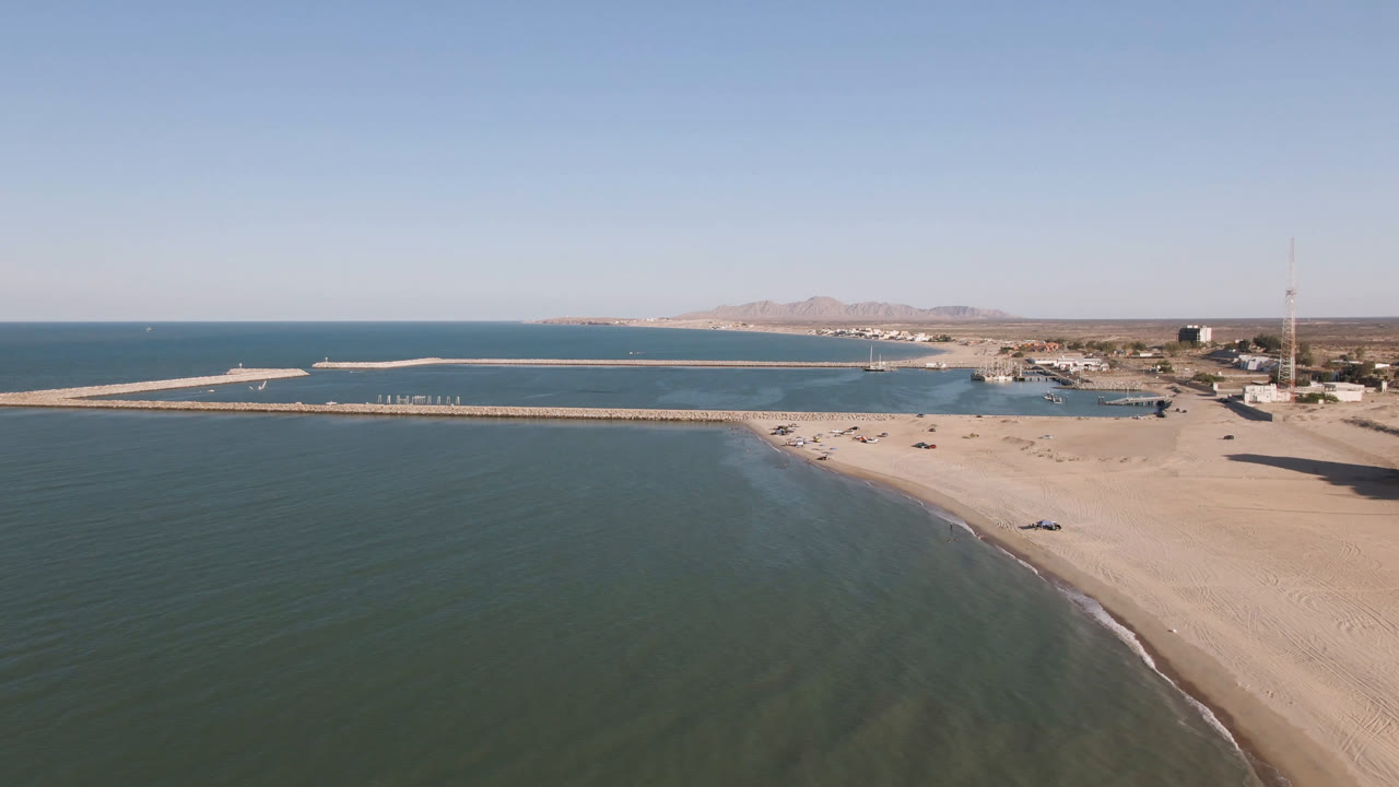 Aerial frame over San Felipe, Baja California, with green-blue water in the foreground, a sandy beach curving right, a long pier across the bay, distant mountains, and no visible people.