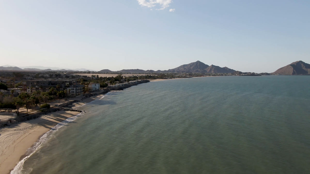 Opening aerial frame over San Felipe, Mexico, with the sandy beach and low buildings on the left, green-blue water across the frame, distant mountains on the horizon, and no visible people.